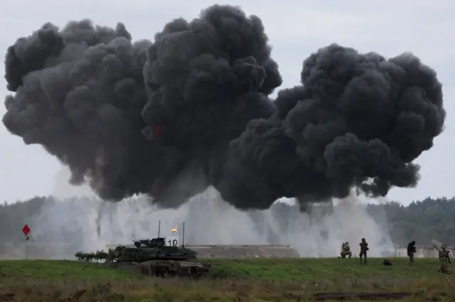 Soldiers operate near Polish Abrams tank as Polish forces and Nato soldiers hold military exercises at a military range in Wierzbiny near Orzysz, Poland, 17 September 2025. A large black cloud of smoke can be seen above the tank, with soldiers silhouetted against lighter grey smoke near the ground.