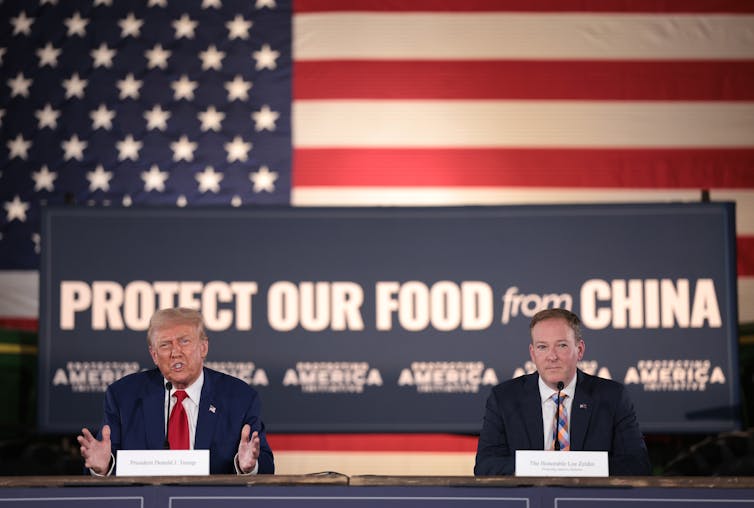 Two men in suits sit at a table in front of a giant American flag and a sign saying 'Protect our food from China.'