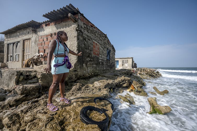 A woman stands outside an old home showing where sea level rise has eroded the shoreline nearly to the home's foundation.