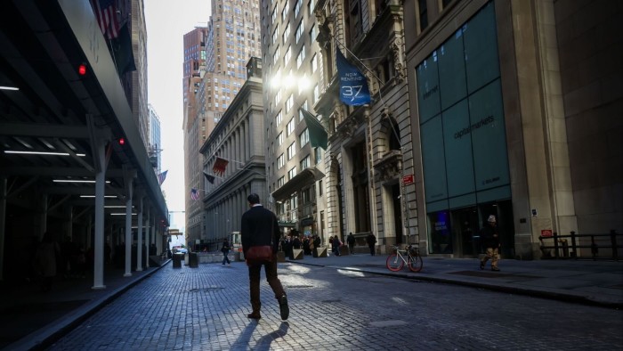 Pedestrians walk along cobblestone Wall Street past historic buildings and flags near the New York Stock Exchange.