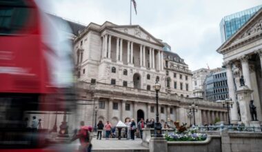 The Bank of England building with people gathered outside and a red bus passing in the foreground.