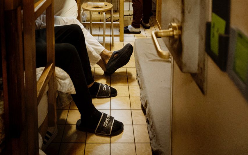 In een krappe en volle cel kijkt een man door een getralied raam naar buiten. Twee andere mannen zitten op een stapelbed, op de grond ligt een matras. 