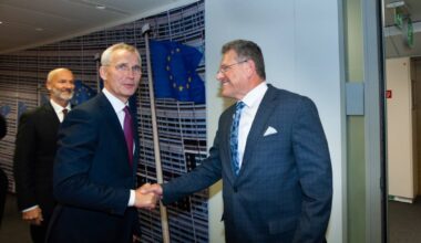 To personer håndhilser i en moderne kontorinnstilling. Europeiske flagg er synlige i bakgrunnen. Det er flere personer i nærheten som observerer situasjonen. Miljøet gir inntrykk av en formell møteplass.