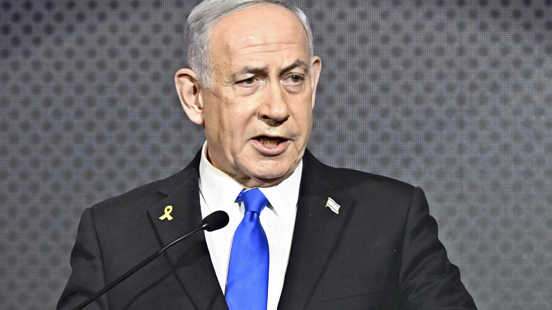 Der israelische Ministerpräsident Benjamin Netanjahu spricht im Anzug vor dunkelgrauem Hintergrund an einem Podium.
