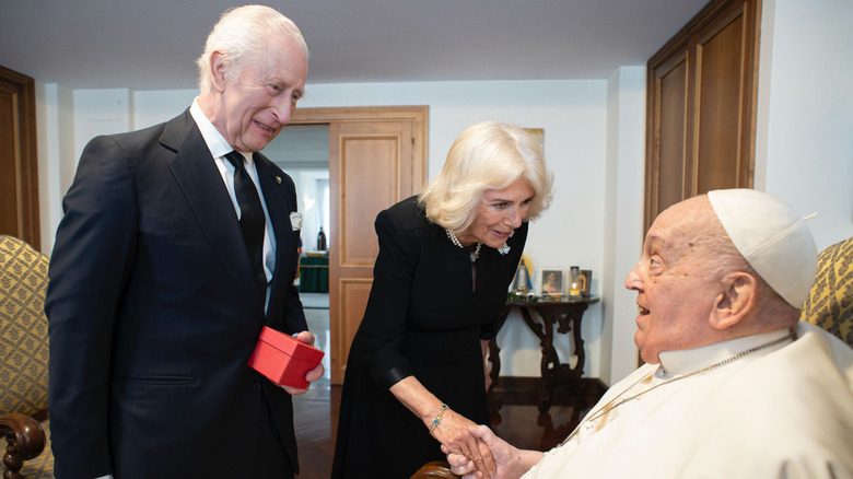 King Charles holding a box while Queen Camilla shakes hands with Pope Francis