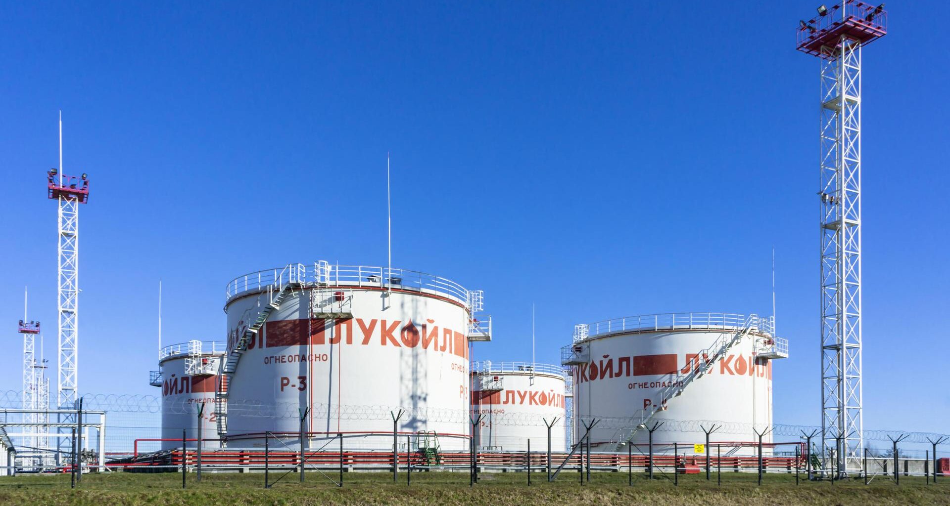 Mehrere umzäunte große weiße Öltanks unter strahlend blauem Himmel. Sie sind aus größerer Entfernung fotografiert.