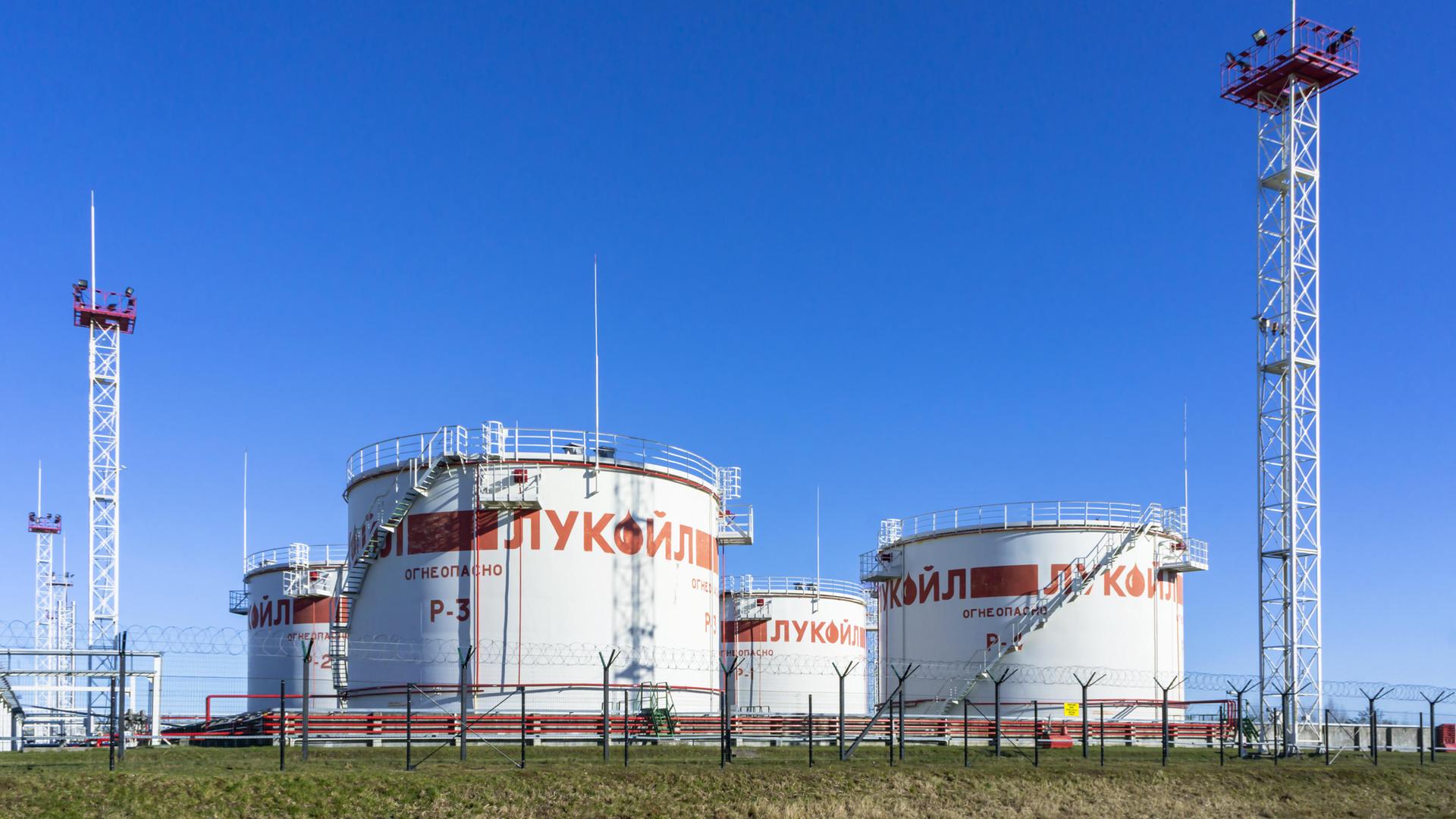 Mehrere umzäunte große weiße Öltanks unter strahlend blauem Himmel. Sie sind aus größerer Entfernung fotografiert.