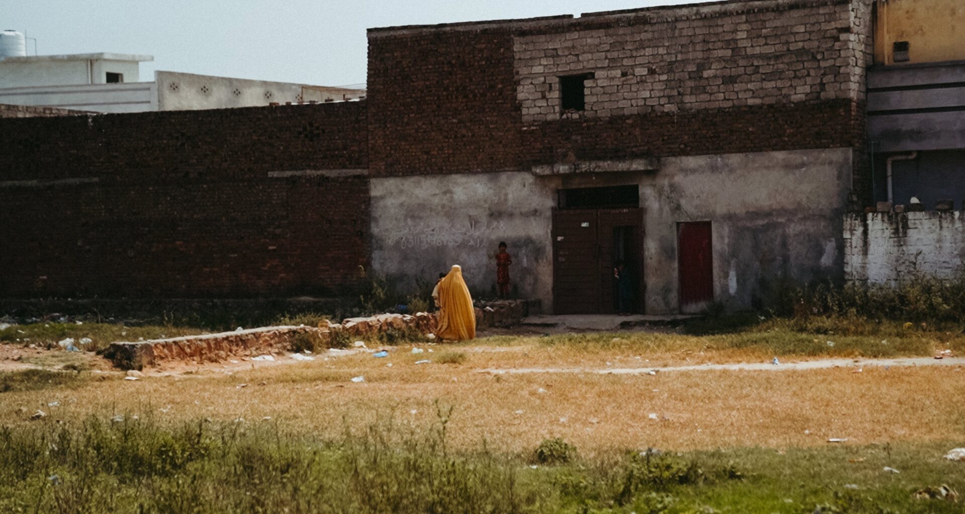 Un homme en habit jaune marche dans un terrain vague, devant des bâtiments en brique.