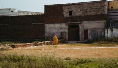 Un homme en habit jaune marche dans un terrain vague, devant des bâtiments en brique.