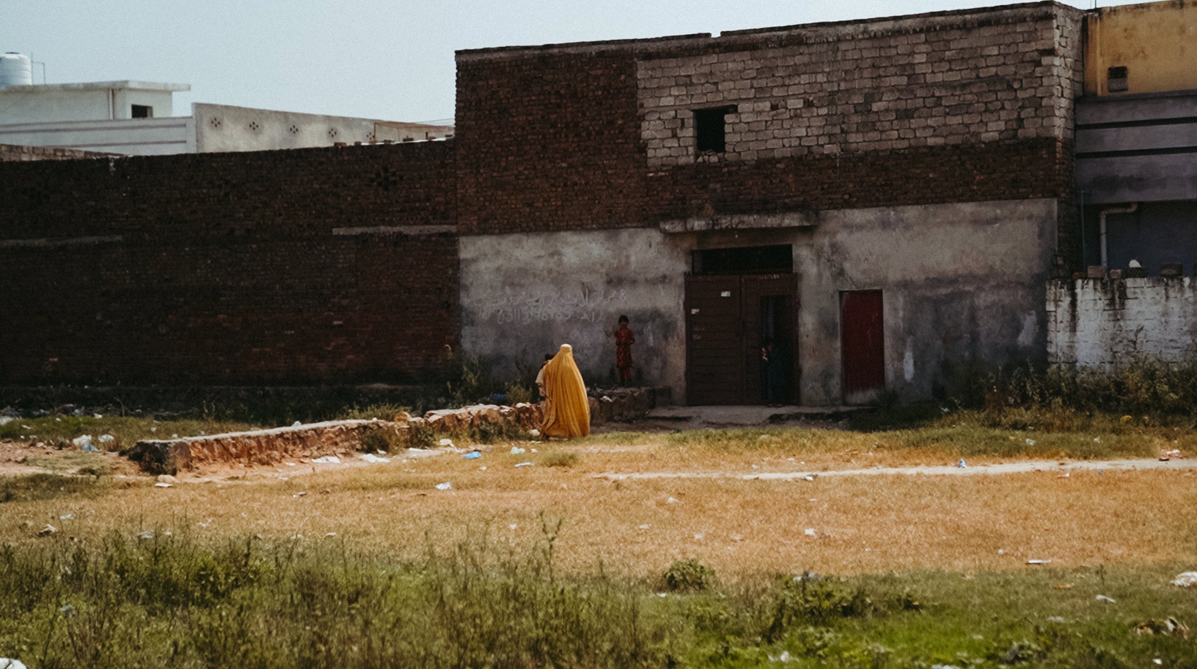 Un homme en habit jaune marche dans un terrain vague, devant des bâtiments en brique.