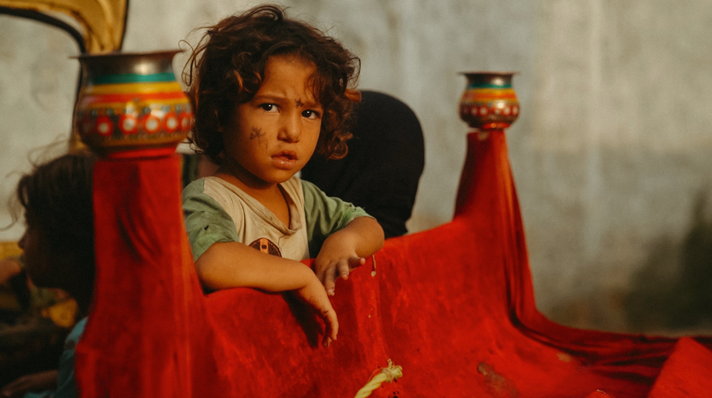 Un enfant aux cheveux bouclés, assis sur une banquette rouge, regarde avec curiosité.
