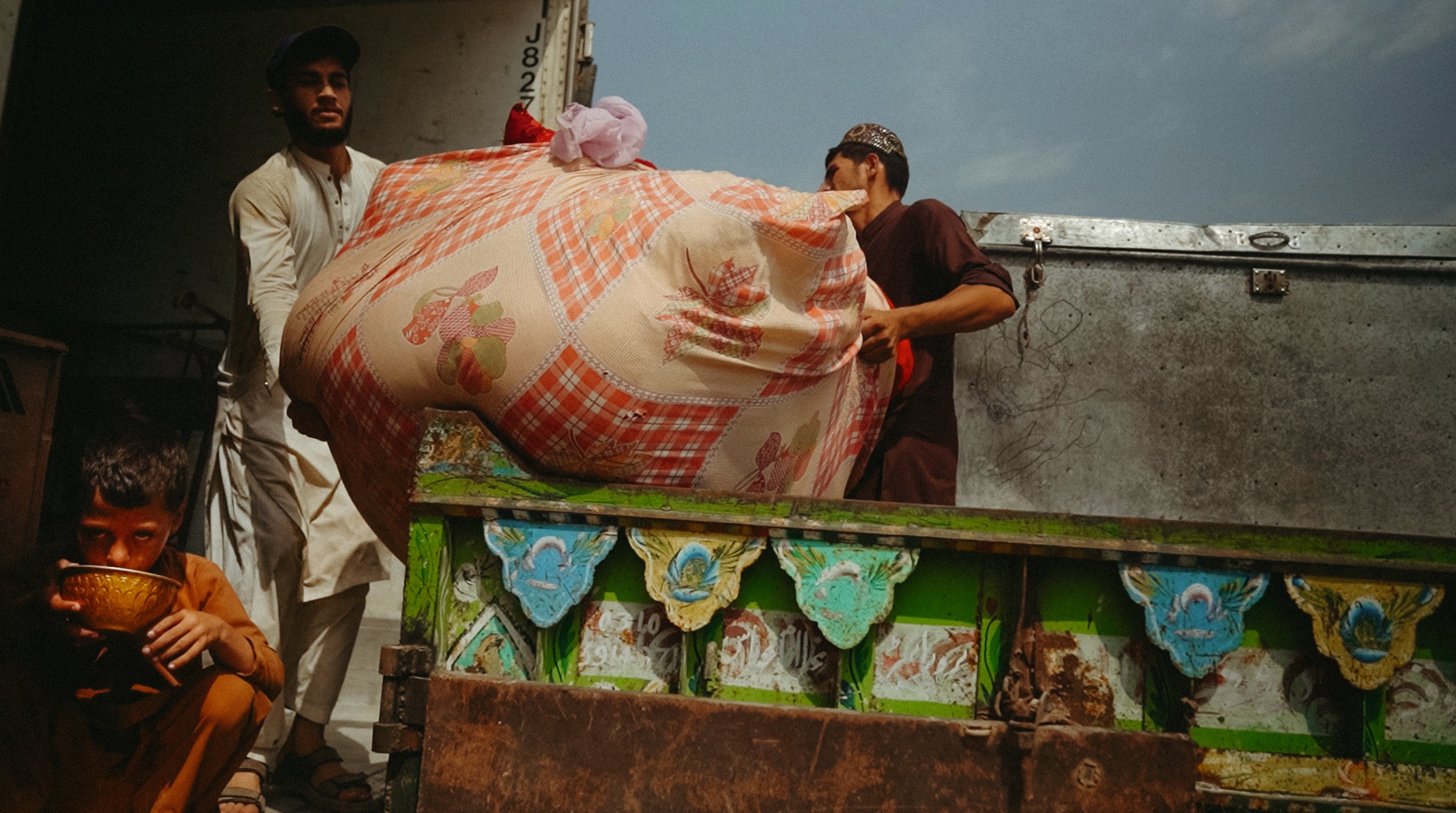 Deux hommes transportent un gros sac, un enfant les observe, ambiance de livraison.