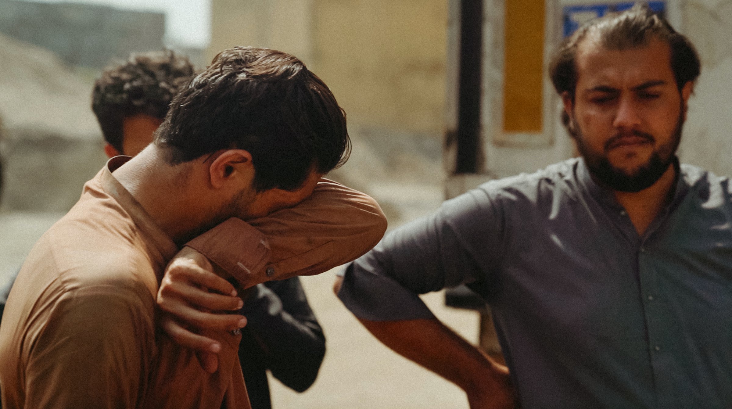 Trois hommes discutent, l'un cachant son visage, dans un cadre extérieur flou.