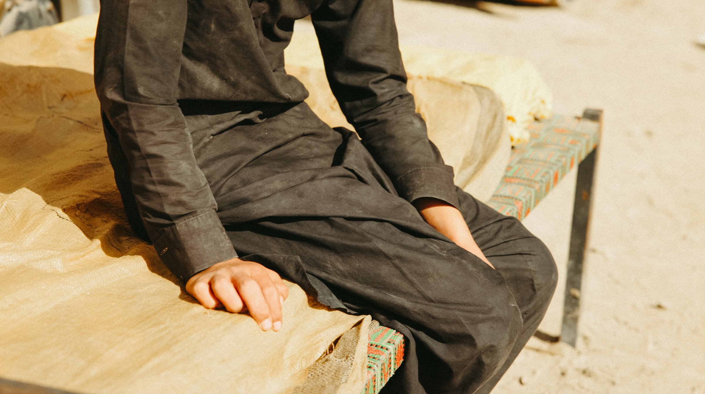 Une personne assise sur un lit, vêtue d'une robe noire, sur un sol sablonneux.