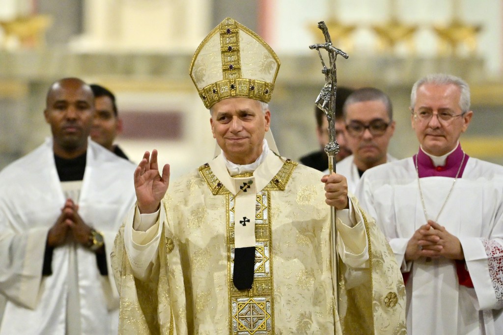 Pope Leo XIV presiding over a mass, wearing a white and gold mitre and vestments, holding a crucifix staff, and raising his right hand in blessing.