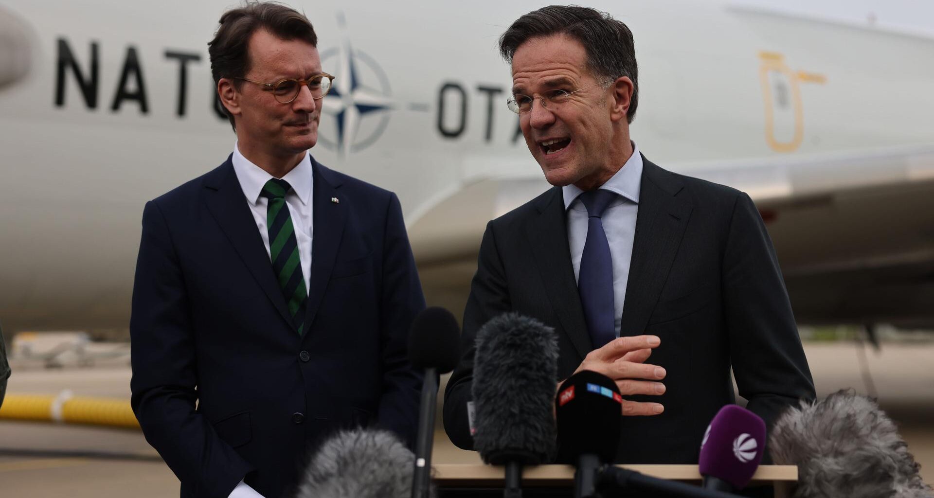 Hendrik Wüst (l, CDU), Ministerpräsident von Nordrhein-Westfalen, Mark Rutte, NATO-Generalsekretär, sprechen vor Mikrofonen, hinter ihnen ist ein Flugzeug der NATO.