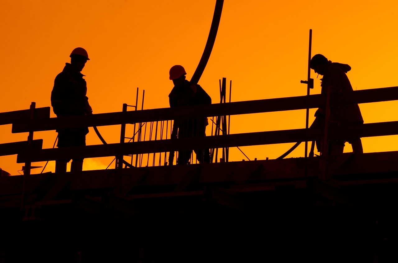 Silhouettes of construction workers on a building framework against an orange sunset sky.
