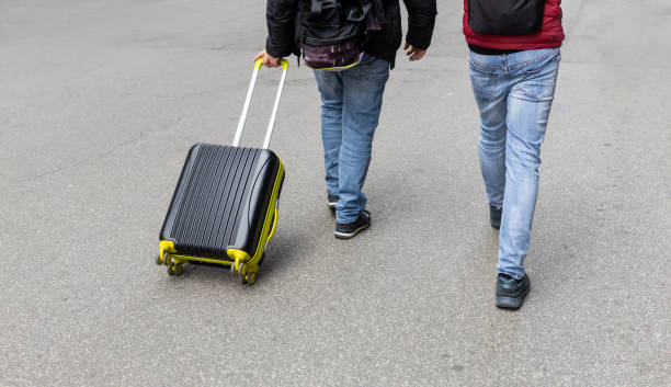 Two travelers walking on a street, one pulling a black suitcase with yellow accents.