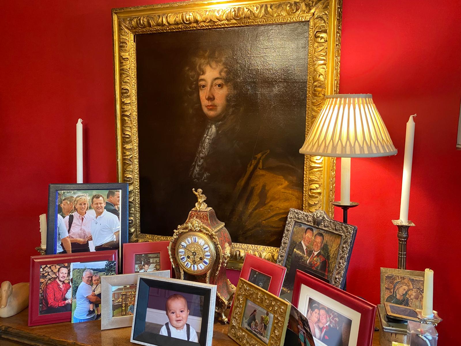 Framed photos, a clock, a lamp, and candles against a red wall beneath a large painting.