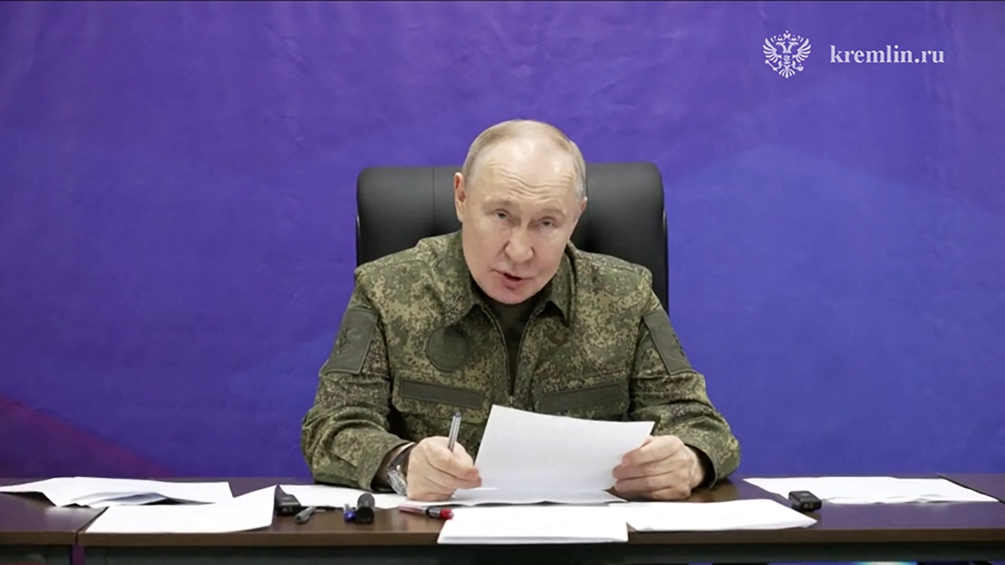 President Putin in military fatigues sits at a desk, speaking, with papers and pens in front of him.