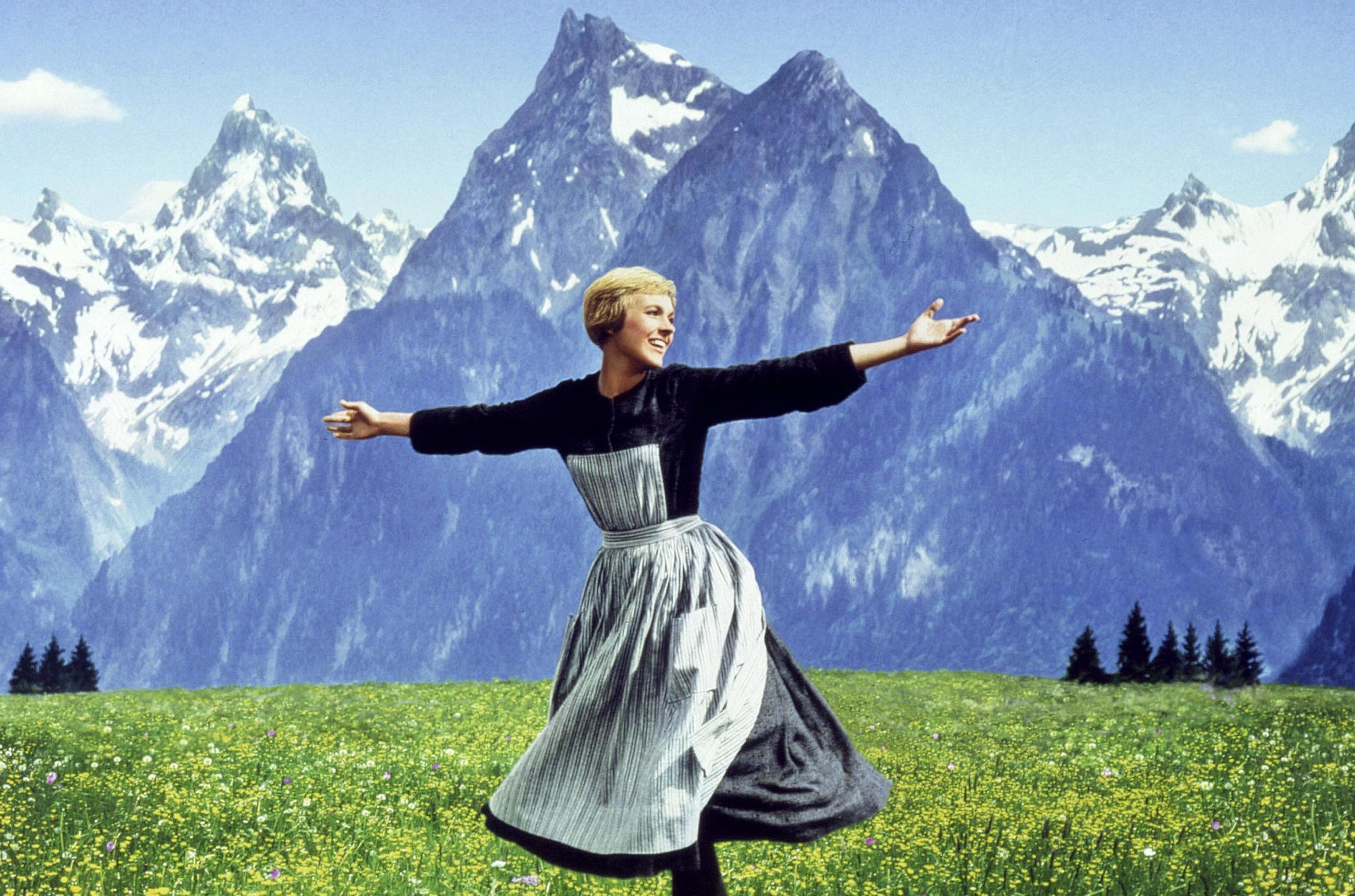 Julie Andrews in The Sound of Music, arms outstretched in a field of yellow flowers with mountains in the background.