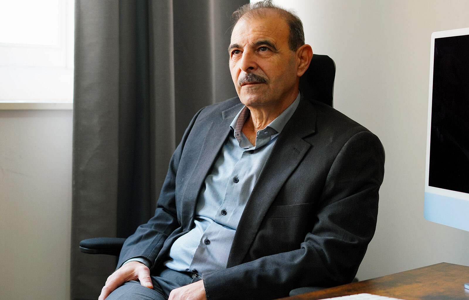 A man in a suit leans back in a chair at his desk, looking contemplatively into the distance. Behind him is a darkened computer monitor. He is Anwar al-Bunni, one of a million Syrian refugees who came to Germany to flee the Assad regime