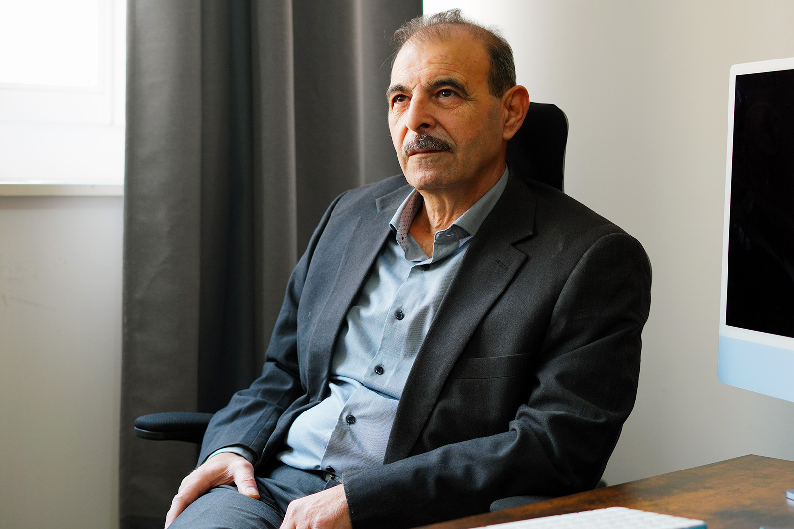 A man in a suit leans back in a chair at his desk, looking contemplatively into the distance. Behind him is a darkened computer monitor. He is Anwar al-Bunni, one of a million Syrian refugees who came to Germany to flee the Assad regime