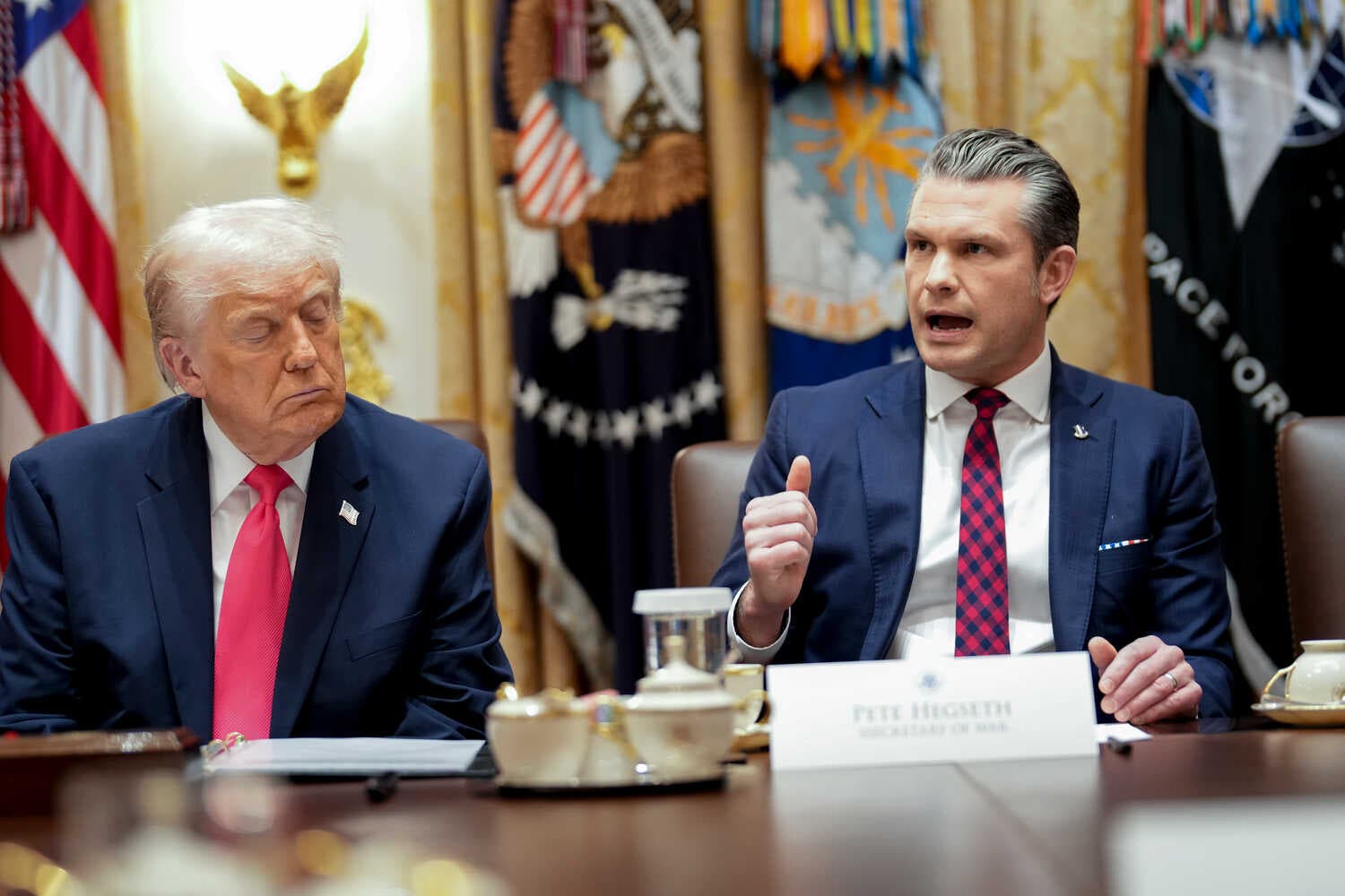 President Trump seated at a table with his eyes closed and his face angled toward Secretary of Defense Pete Hegseth, who is seated next to him and speaking.