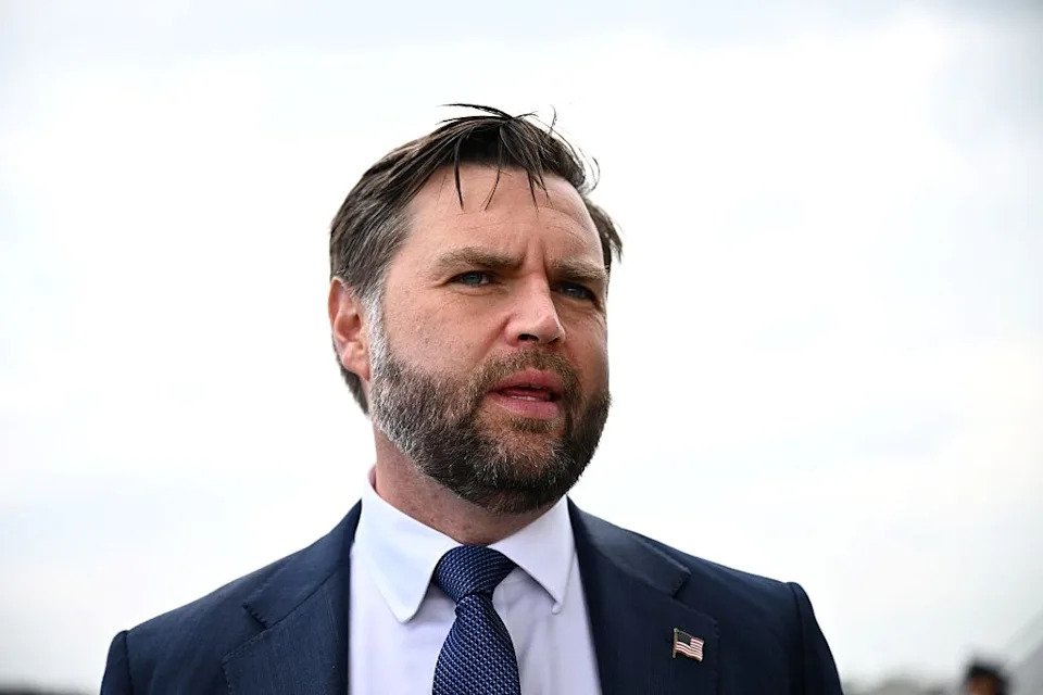 jd vance in a suit with a U.S. flag lapel pin looking ahead during an outdoor event