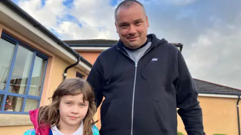 a man and a little girl are pictured outside a school building at Bready primary School. The little girl is on the left, and has long brown hair.  The man is wearing a blue hoodie xipped up