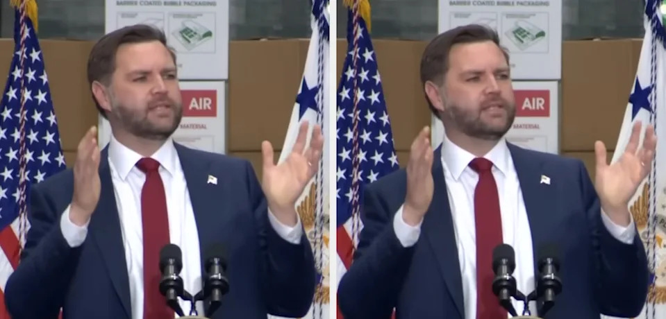 A man in a suit and red tie gestures while speaking at a podium with flags in the background