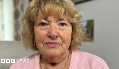 Joyce Gifford with blue eyes and golden-coloured, short, wavy hair with a fringe. She is wearing a baby pink wool jumper with a thin, gold necklace and thin, gold, hoop earrings.