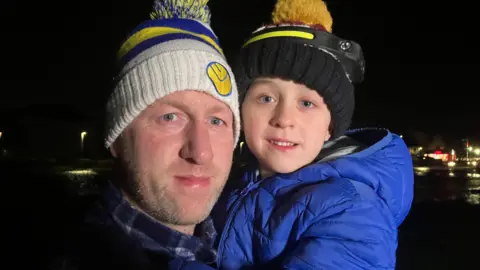 Stephen O'Flynn is holding Rían Patton O'Flynn in his arms and both are smiling. They are both wearing wooly bobble hats and large coats. 
