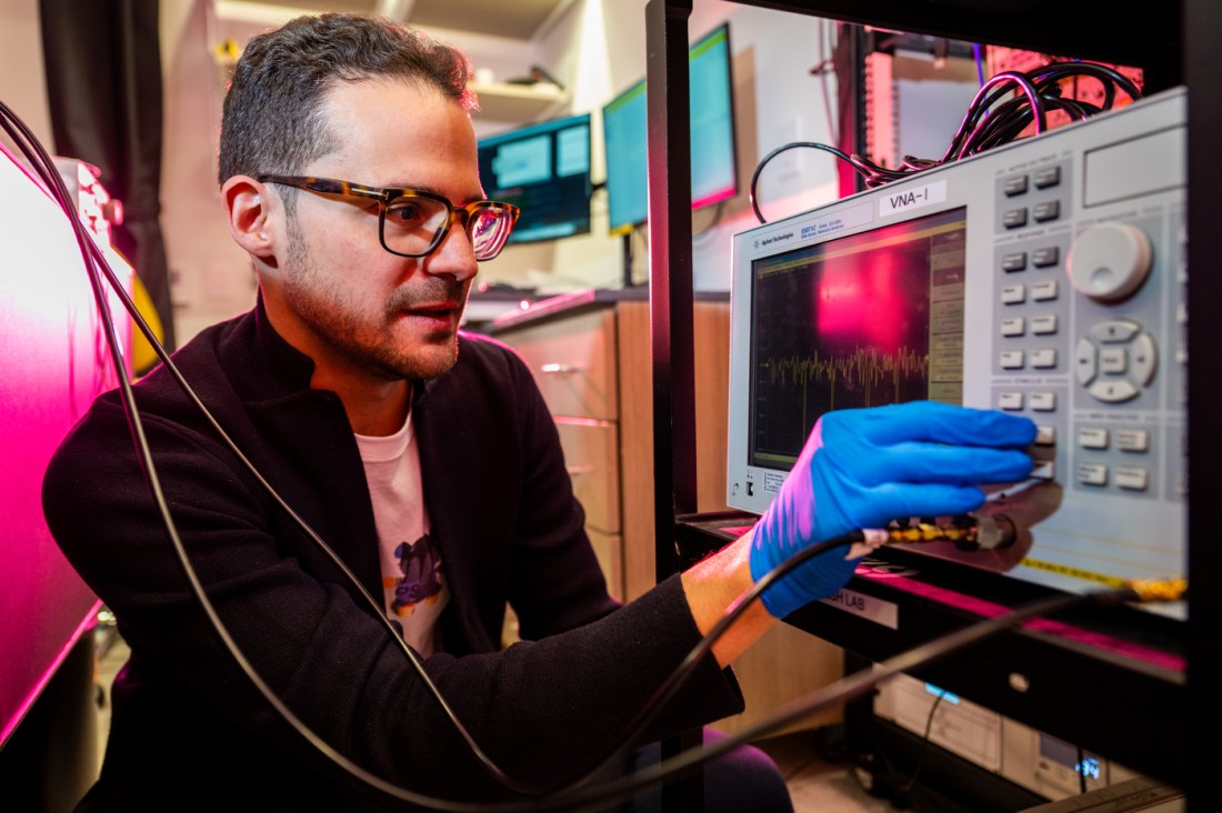 A man with glasses and blue latex gloves crouches next to a device providing an oscilloscope-like display of a line with peaks and valleys.