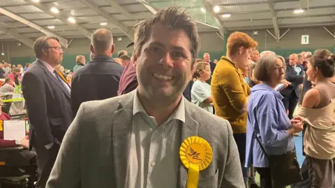 BBC Leigh Frost wearing a grey blazer with a white striped shirt. He also has a yellow rosette on his left side with the Liberal Democrat bird logo on it. He has short brown hair and is stood in front of a crowd of people at the count.
