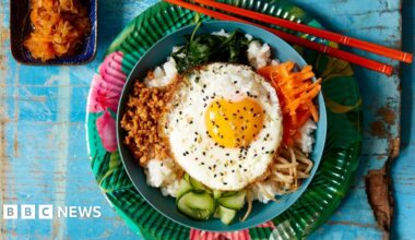 Bibimbap, a Korean dish featuring rice and various vegetables such as carrots, spinach, bean sprouts and a fried egg on top, served with chopsticks, and a side dish of kimchi.
