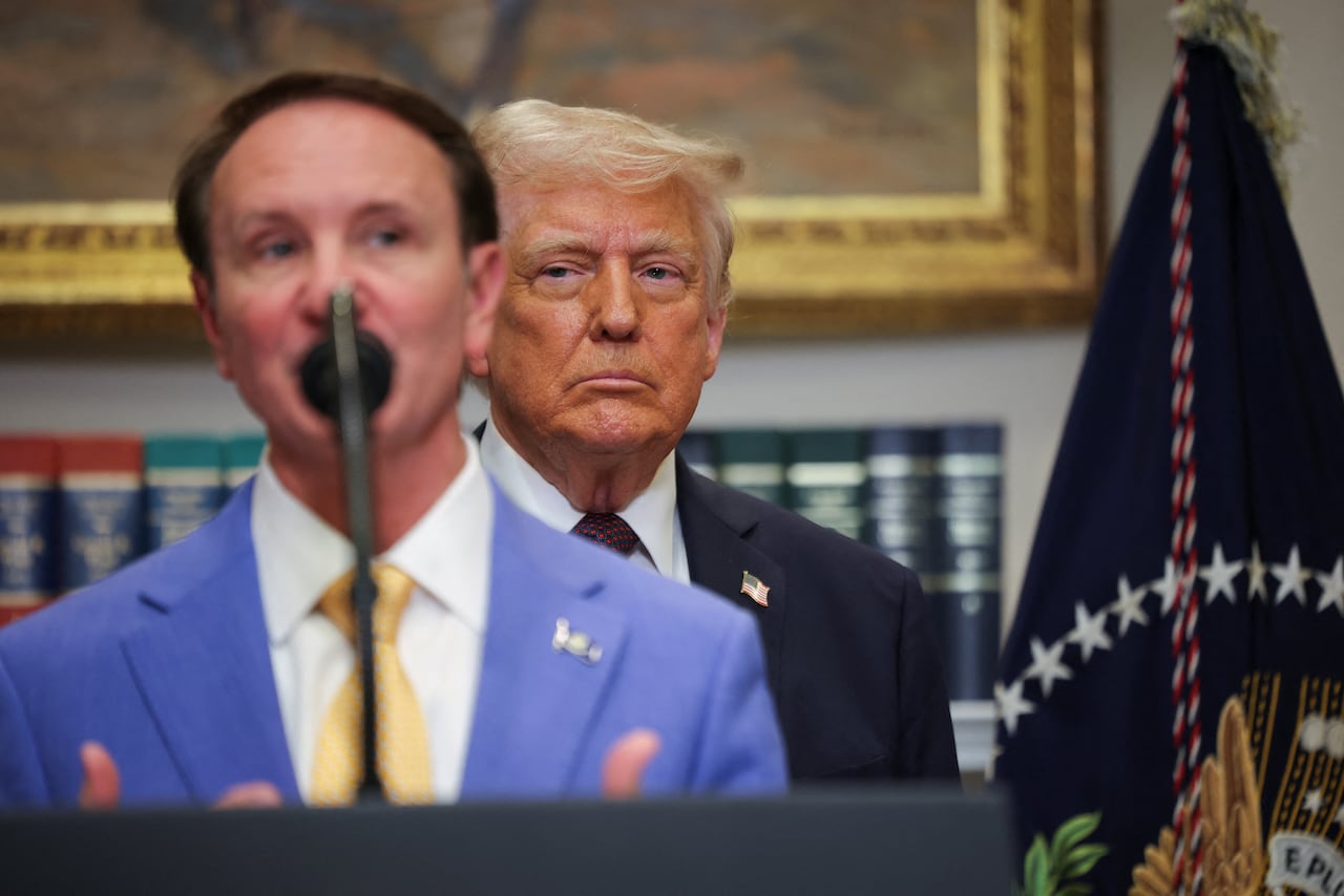 A man speaks while standing in front of a microphone, while U.S. President Donald Trump looks on from the background