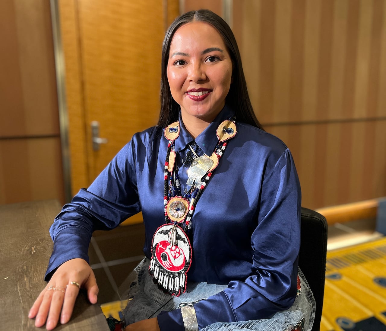 A woman with long black hair, wearing a blue blouse and a beaded necklace, sits at a table.