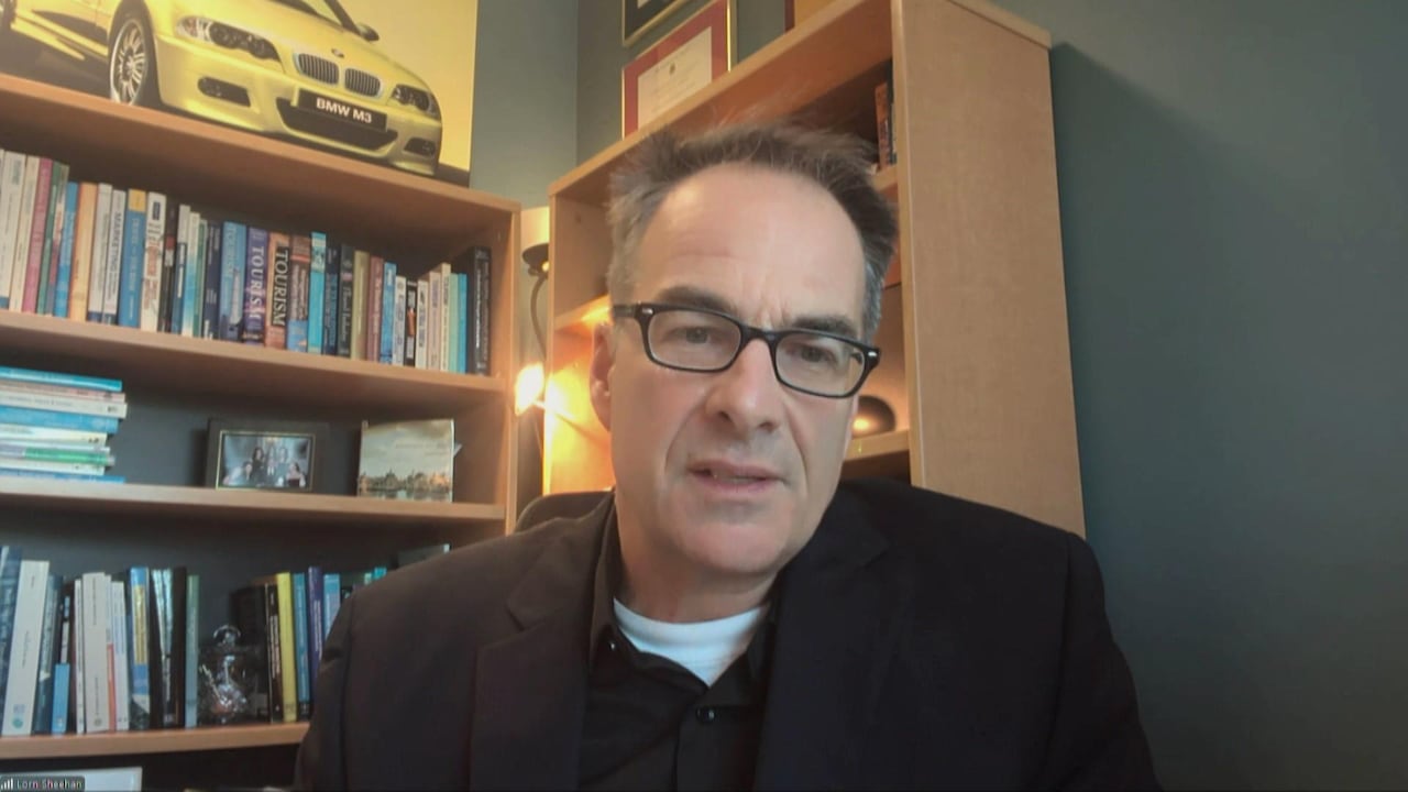  A man in a dark suit jacket sits in front of his computer staring into the screen. Bookshelves line the walls behind him.