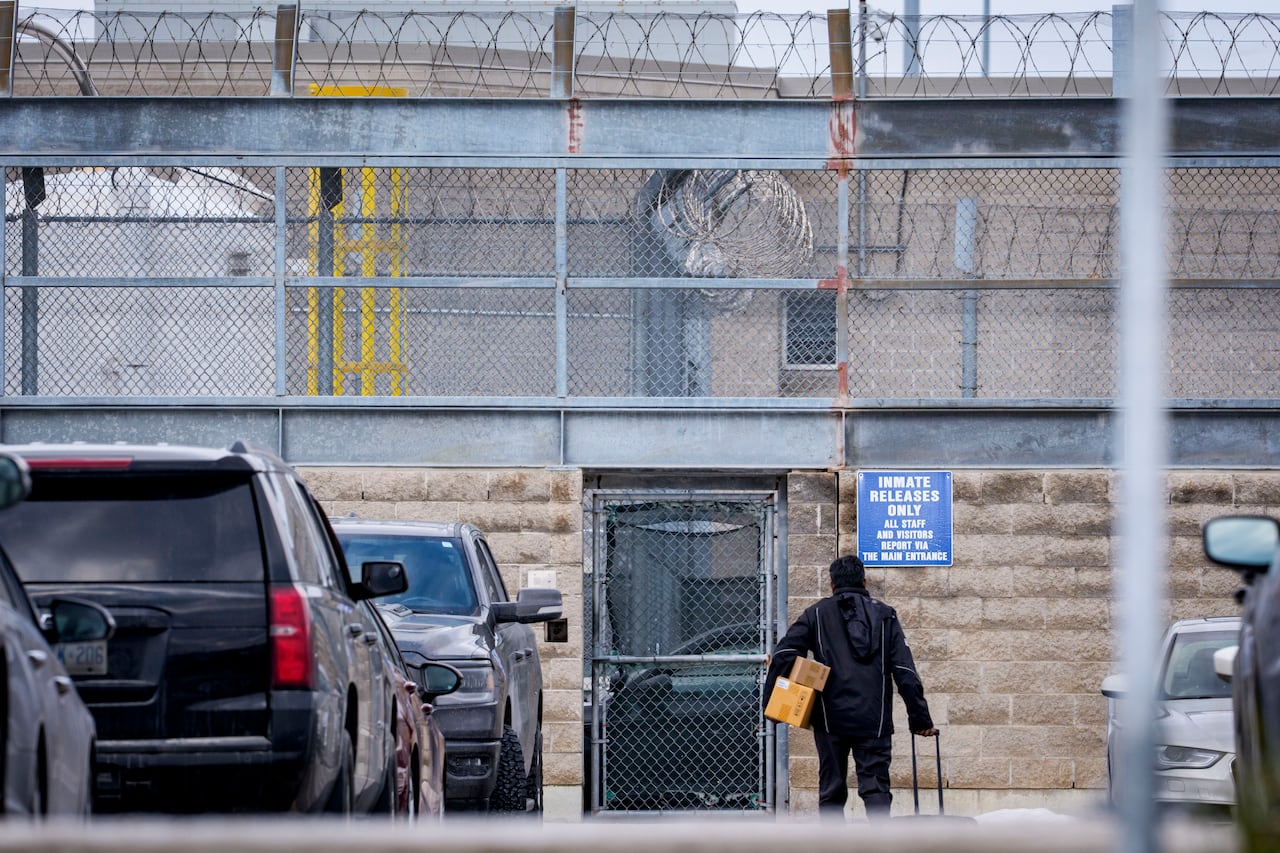 A person holding two small boxes pulls a wheeled bag while walking towards a gate of a women's prison, where a sign on the wall reads, "Inmate releases only."