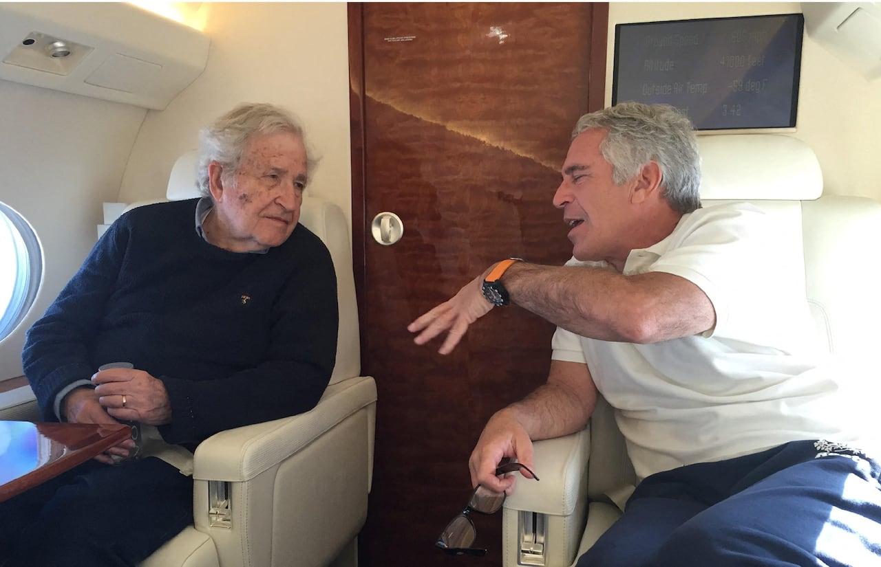Noam Chomsky, with grey hair and wearing a dark sweater, sits in a plane seat on the left, looking at Jeffrey Epstein, with grey hair and wearing a white polo t-shirt and blue pants, seated on the right, talking and gesturing with his left hand.
