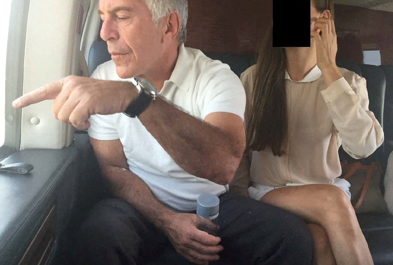 A photograph of Jeffrey Epstein, with grey hair and waring a white polo t-shirt and grey pants, sitting on a plane and pointing out the window. A woman in a beige dress, with long, dark hair, sits behind. Her face is redacted with a black box.