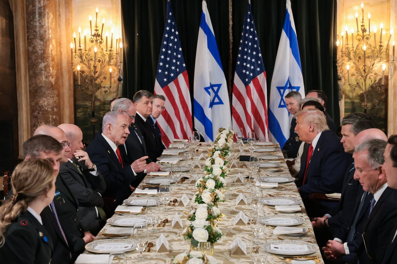 A long table with about 24 people seated around it, US and Israeli flags at the head