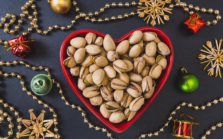 Roasted salted pistachios shelled in heart shaped red bowl on black background with Christmas decorations around the bowl.