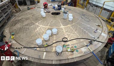 Workers in red protective clothing inspect and work on a large circular metal structure inside an industrial workshop. The structure has a patterned surface with a central opening and several white buckets placed on top. Tools, cables, and equipment are scattered around.