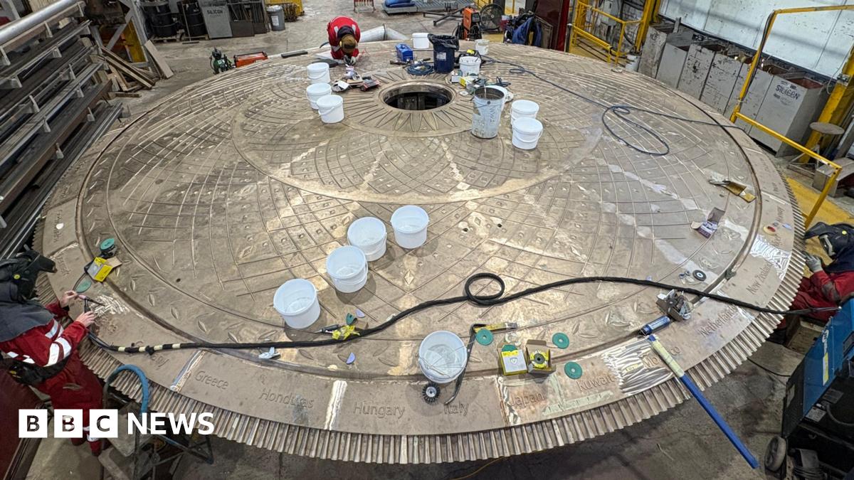 Workers in red protective clothing inspect and work on a large circular metal structure inside an industrial workshop. The structure has a patterned surface with a central opening and several white buckets placed on top. Tools, cables, and equipment are scattered around.