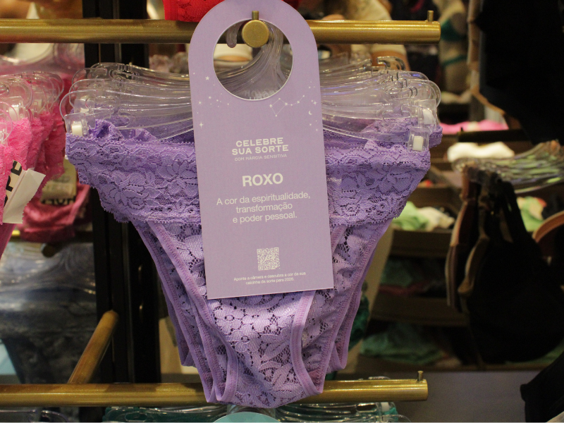 Calcinha roxa de renda exposta em um suporte com tag informativa sobre espiritualidade e poder pessoal.