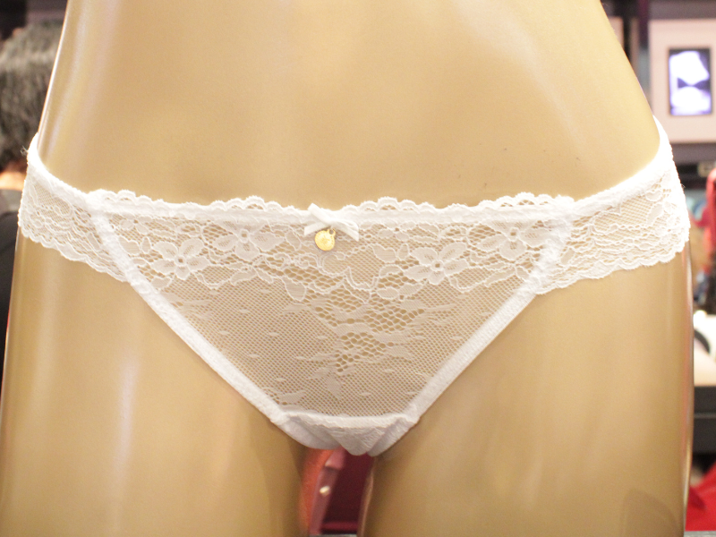 Calcinha branca de renda transparente com um pequeno laço e pingente dourado central, exibida em manequim.