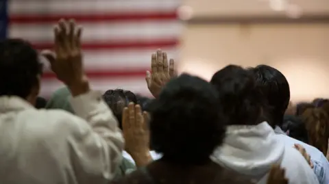 Getty Images A shot of the back of people's heads as they stand and take the oath of citizenship in front of an American flag