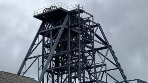 South Crofty tin mine. A grey metal structure is suspended above the ground with ladders/steps leading to the very top. The sky is also grey.